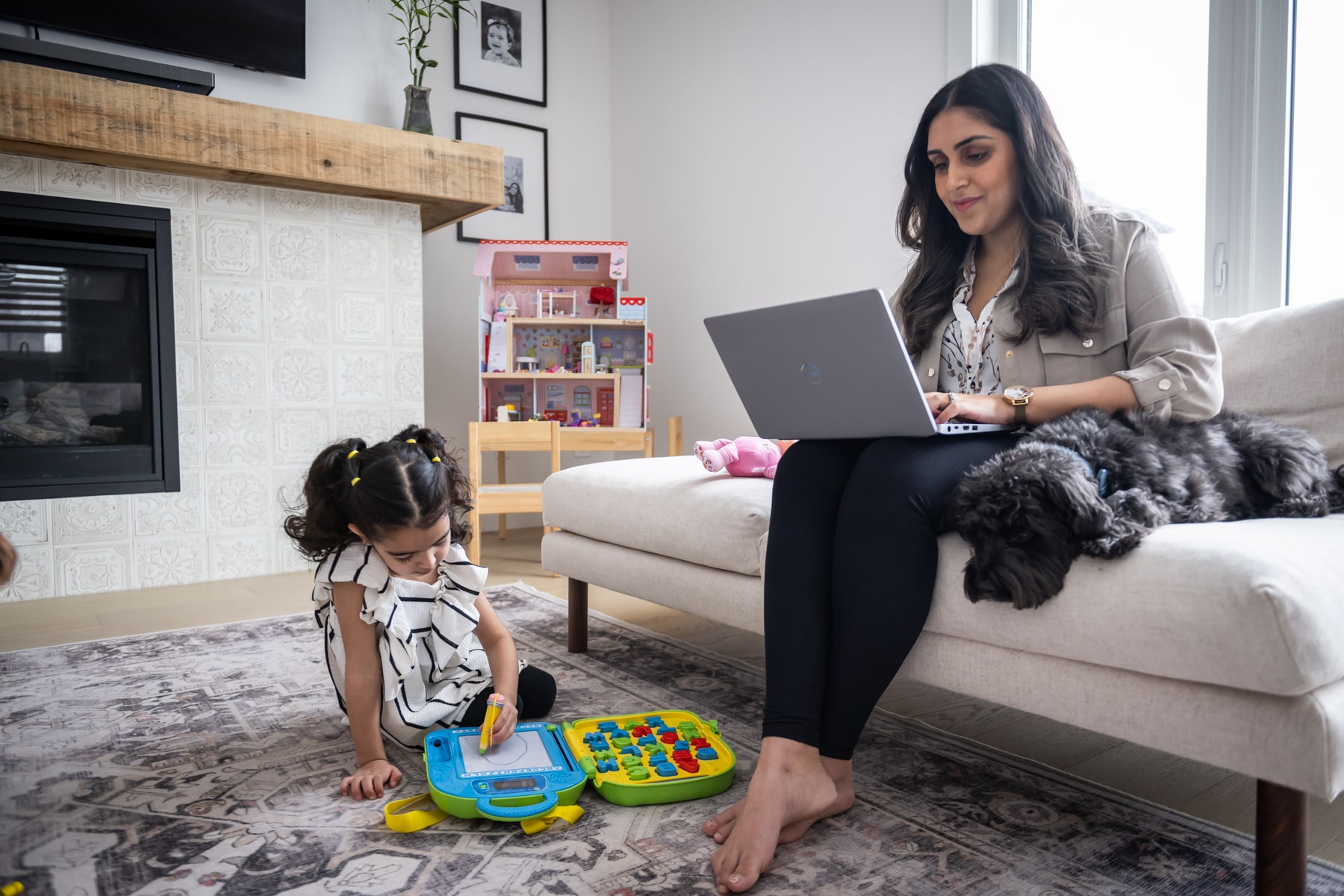 mother sits on couch studying on laptop while child plays nearby