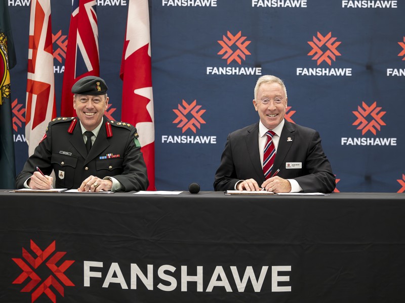 Colonel Chris Brown and President Peter Devlin seated at a table in front of Fanshawe backdrop to sign an MOU agreement.